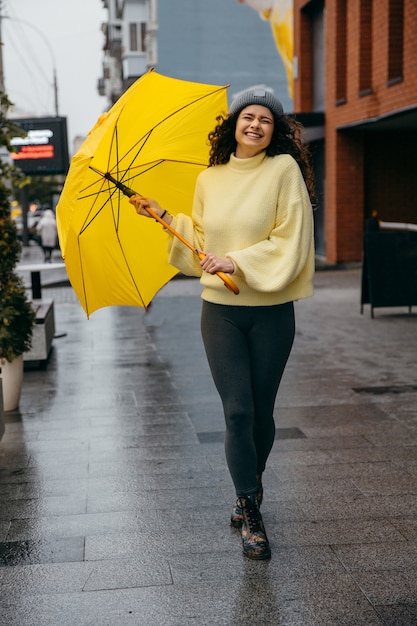 Charmante jeune femme bouclée utilise un parapluie jaune dans la rue de la ville de mégapole en jour de pluie