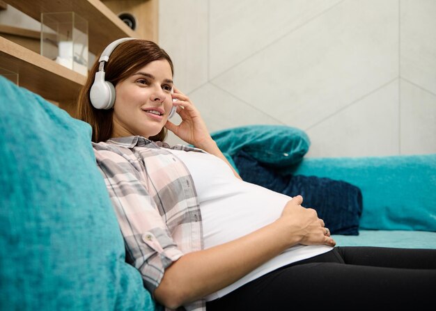 Charmante jeune femme caucasienne rousse enceinte reposant sur un congé de maternité caressant doucement son ventre de femme enceinte et appréciant écouter de la musique classique sur des écouteurs à la maison allongée sur le canapé