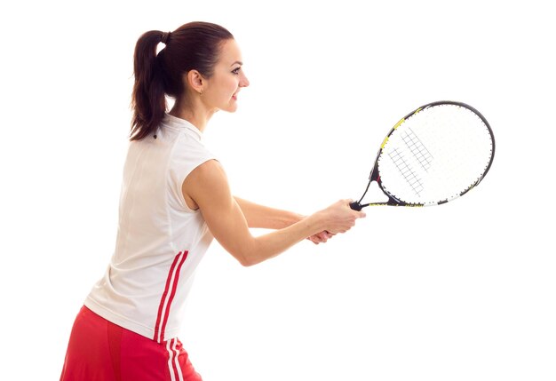 Charmante jeune femme en chemise de sport blanche et jupe rouge avec queue de cheval foncée tenant une raquette de tennis