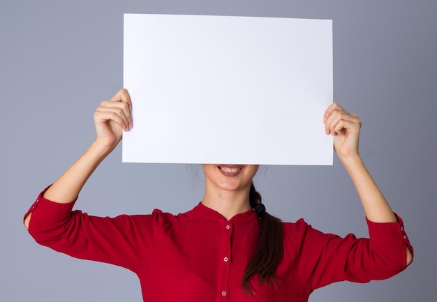 Charmante jeune femme en chemisier rouge tenant une feuille de papier blanche sur fond gris en studio