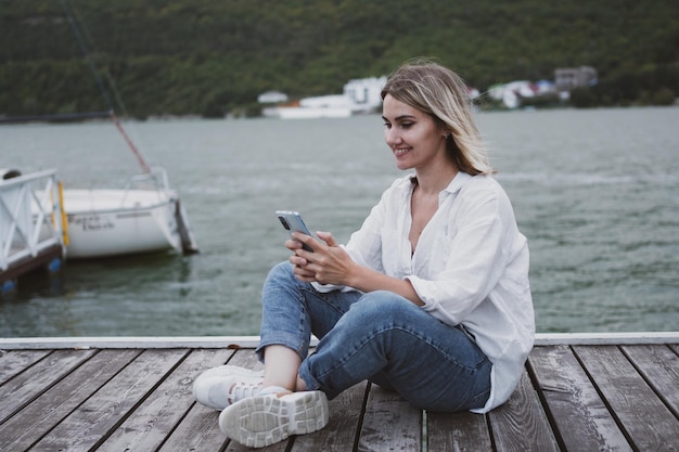 Charmante jeune femme est assise sur la jetée sur le magnifique lac de montagne et communique sur téléphone mobile