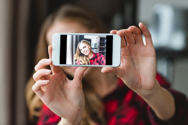 Charmante jeune femme joyeuse en chemise à carreaux prenant selfie avec smartphone