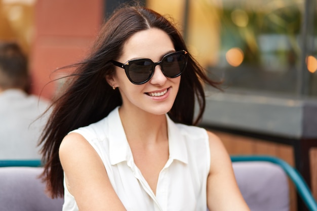 Charmante jeune femme à la mode shads, chemisier blanc, se trouve au café en plein air contre floue