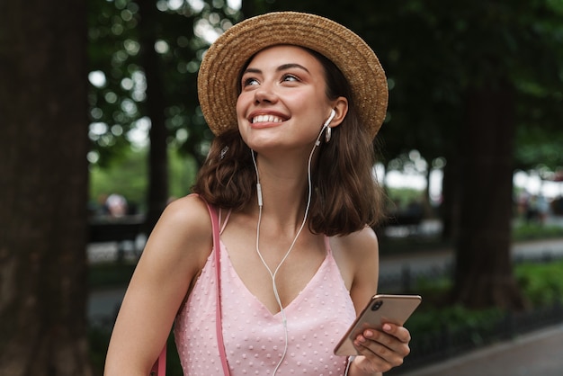 charmante jeune femme portant un chapeau de paille utilisant un téléphone portable et des écouteurs tout en marchant dans un parc verdoyant