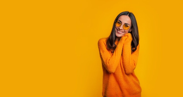 Charmante jeune femme en pull jaune et lunettes de soleil alors qu'elle pose avec le sourire sur fond jaune et regarde la caméra