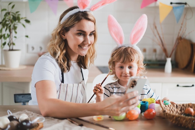 Charmante jeune femme et son fils de deux ans dans des oreilles de lapin peignent des oeufs de Pâques avec des peintures assis à la table de la cuisine