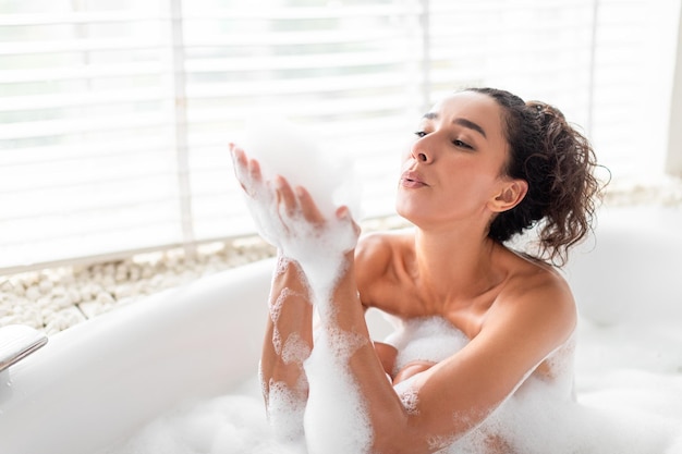 Charmante jeune femme soufflant des bulles de savon tout en se relaxant dans un bain à la maison espace libre