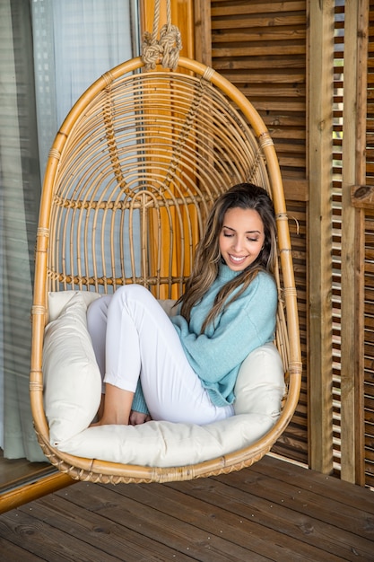 Charmante jeune femme souriante en vêtements blancs et bleus assis sur un confortable fauteuil suspendu en osier en rotin tout en se détendant sur la terrasse de la maison