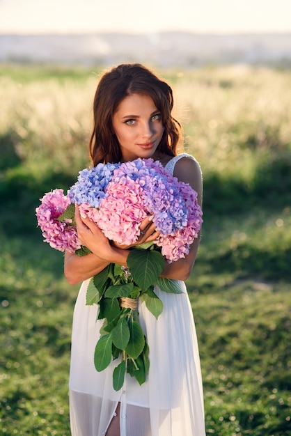 Une charmante jeune fille avec un beau sourire dans une robe blanche avec un bouquet de fleurs aux couleurs tendres au coucher du soleil.