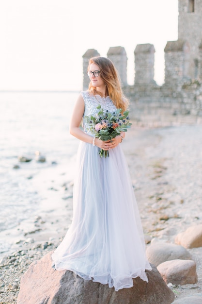 Une charmante jeune fille dans une robe de mariée bleue debout sur des rochers près du château de Rocca Scaligera à Sirmione. Lac de garde