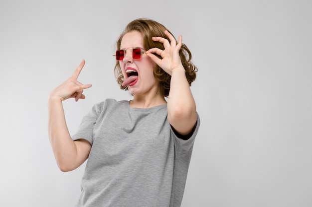 Charmante jeune fille dans un t-shirt gris sur fond gris. Fille à lunettes rouges aux yeux carrés. La jeune fille montre un bouc bascule