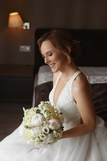 Une charmante jeune mariée heureuse en robe de mariée élégante posant avec un bouquet de mariée à l'intérieur