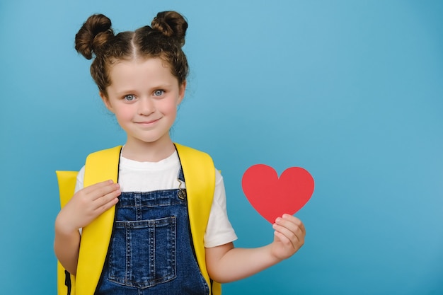 Charmante jolie écolière mignonne avec un sac à dos jaune tenant un coeur de papier rouge à la main, heureuse de regarder la caméra, posant isolée sur un mur de fond bleu studio. Émotions des gens et concept de retour à l'école