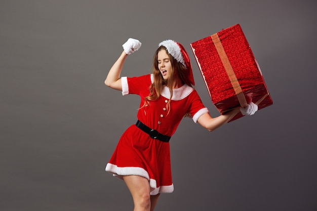 Charmante Mme Claus vêtue de la robe rouge, du bonnet du Père Noël et des gants blancs tient dans sa main l'énorme cadeau de Noël sur fond gris .