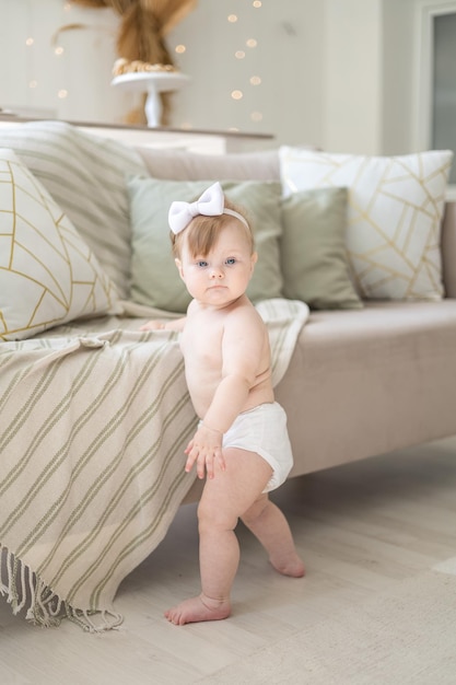 Une charmante petite fille en couches se tient près du canapé à la maison dans un salon lumineux avec un support que l'enfant apprend à faire les premiers pas