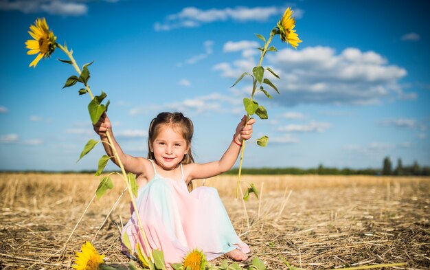 Charmante petite fille dans une longue robe d'été est assise sur un champ sur le fond d'un ciel bleu et nuages blancs