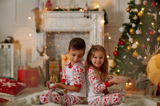Charmante petite fille et garçon en pyjama assis sur un plaid avec des cadeaux et un sapin de noël près de blowi...