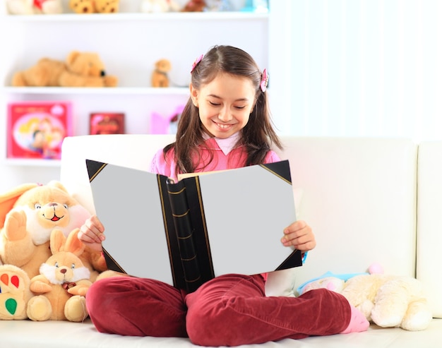 Charmante petite fille lisant un livre avec son jouet sur un canapé blanc