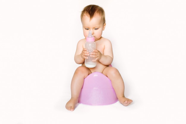 Charmante photo de jolie petite fille tenant une bouteille avec du lait pour bébé et assise sur un pot rose. Isolé sur fond blanc