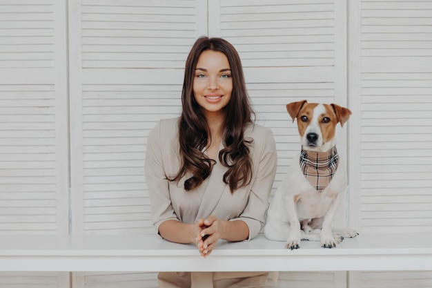 Charmante superbe femme européenne aux cheveux longs noirs regarde directement la caméra garde les mains ensemble mignon chien de race pose près de jouer ensemble ont une relation amicale Propriétaire de l'animal à l'intérieur