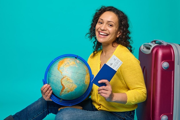 Une charmante voyageuse en pull jaune est assise à côté des bagages, détient un globe, une carte d'embarquement, un billet et un passeport, sourit à pleines dents en regardant la caméra. Concept de tourisme sur fond bleu, espace de copie