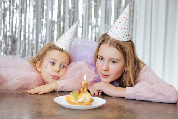 Charmantes petites filles aux cheveux blonds avec des robes de festival lilas et roses et des chapeaux de fête soufflant une bougie sur un gâteau d'anniversaire, assises à l'intérieur éventuellement dans un café avec un fond décoré de paillettes argentées