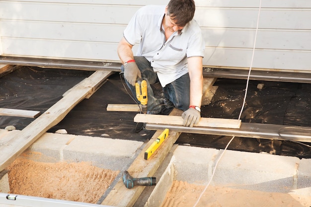 Charpentier de beau jeune homme installant une terrasse extérieure de plancher de bois dans un nouveau chantier de construction de maison