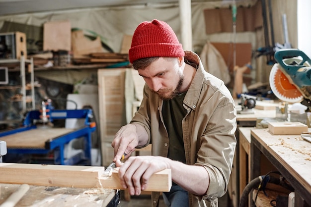 Charpentier caucasien barbu portant une casquette en tricot rouge coupant la mortaise dans une planche de bois à l'aide d'un ciseau