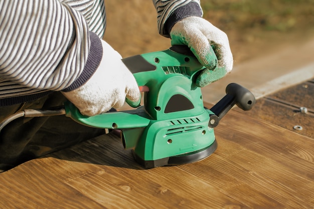 Photo charpentier dans des gants de protection est en train de broyer des planches brûlées avec une ponceuse électrique