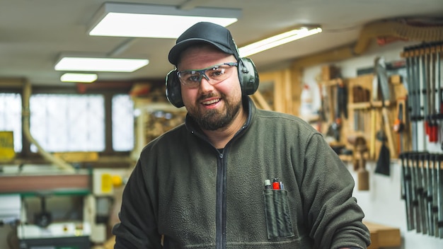 Charpentier masculin prêt à travailler dans des lunettes de protection et des écouteurs souriant à l'intérieur de la caméra