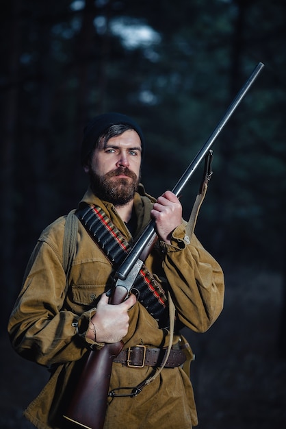 Photo chasseur d'homme barbu brutal sévère dans un chapeau noir et une veste kaki dans un long manteau tient une arme à feu dans sa main dirigée vers la forêt