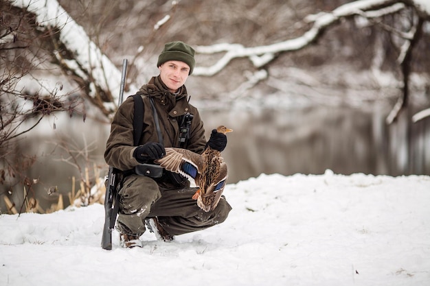 Chasseur mâle en tenue de camouflage armé d'un fusil debout dans une forêt d'hiver enneigée avec des proies de canard