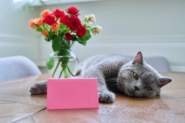 Chat heureux détendu allongé sur la table avec bouquet de fleurs dans un vase et carte vierge avec espace de copie Décoration de vacances invitation félicitations concept cadeau