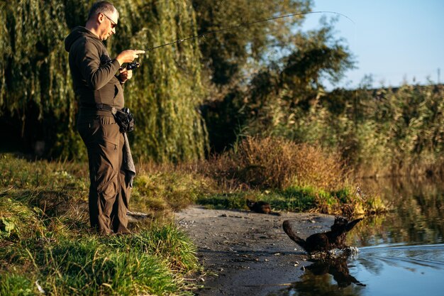 Un chat noir affamé sans-abri attrapant du poisson avec un pêcheur sur la rive de la rivière Un chat noir affamé égaré obtient