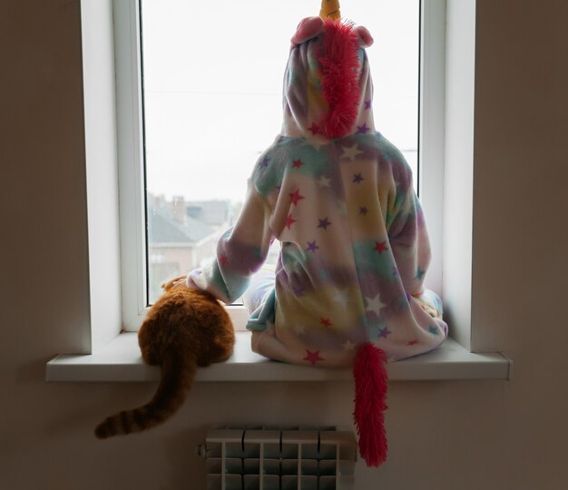 Un chaton au gingembre et une petite fille sont assis sur le rebord de la fenêtre et regardent par la fenêtre.