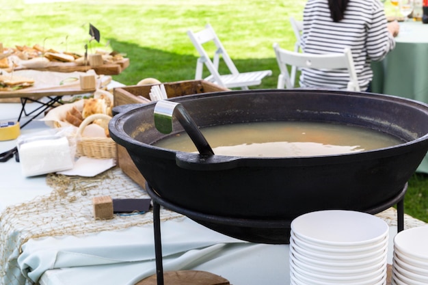 Un chaudron avec de la soupe de poisson se dresse sur une table de buffet sur une terrasse d'été La soupe est servie dans des bols en papier jetables photo horizontale