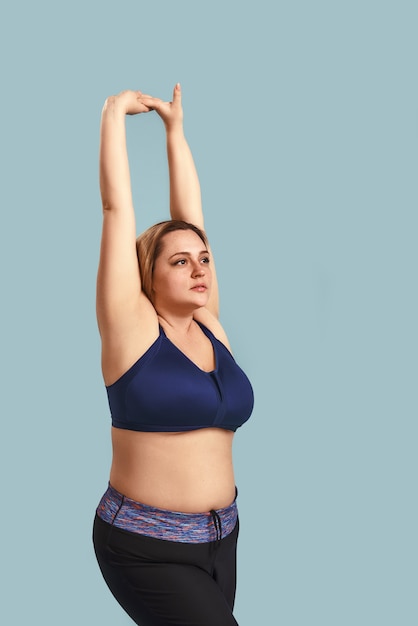Échauffement. Photo verticale d'une jeune femme de taille plus en vêtements de sport faisant des exercices d'étirement à l'intérieur contre un mur bleu. Mode de vie sain. Notion de sport. Perte de poids