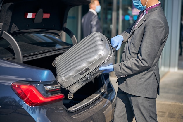 Un chauffeur en costume emballe des bagages dans une voiture près du terminal de l'aéroport tout en portant un masque et des gants de protection