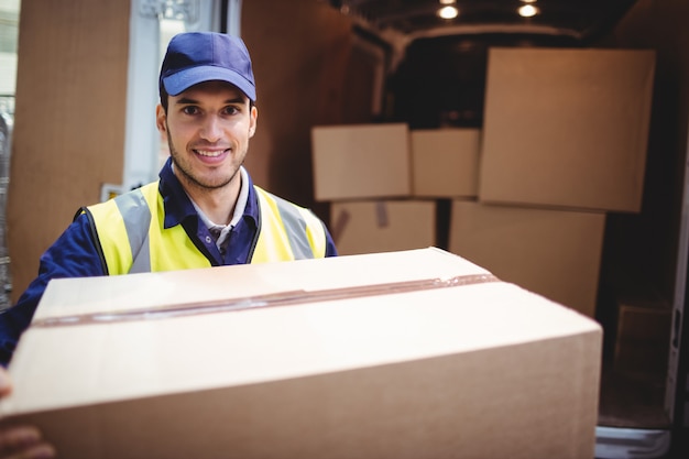 Chauffeur-livreur souriant à la caméra devant son camion transportant un colis devant l'entrepôt