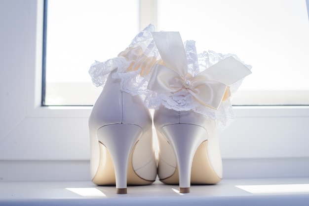 Photo chaussures de mariage blanches de la mariée et de la jarretière par la fenêtre