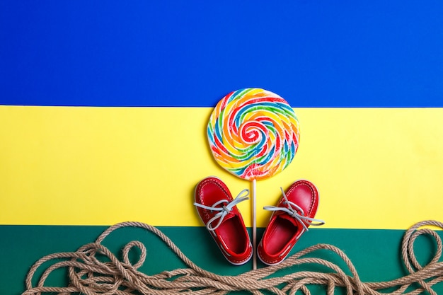 Chaussures de petit bateau rouge près de grosse sucette multicolore et corde sur fond coloré.