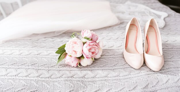 Chaussures à talons hauts de la mariée. Superbe robe de mariée blanche étonnante et chaussures à talons hauts allongés sur le lit près du bouquet de mariée rose dans la chambre