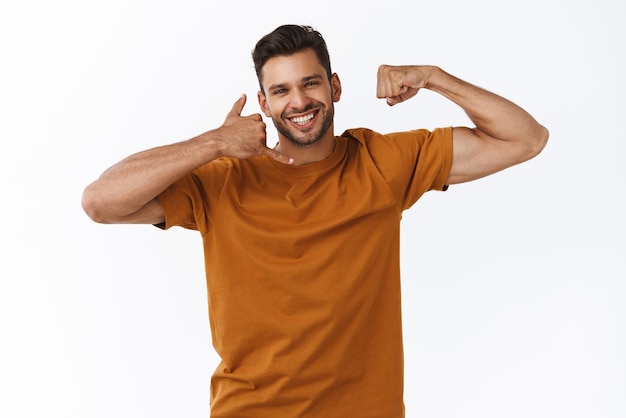 Cheeky goodlooking instructeur de fitness masculin barbu montrant sa force corporelle et faire un geste téléphonique près de l'oreille souriant coquette rire encourager l'appeler si vous voulez gagner en force et faire de l'exercice