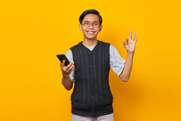 Cheerful asian man holding smartphone et gesticulant signe ok sur fond jaune