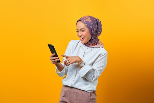 Cheerful Asian woman looking at smartphone obtenir de bonnes nouvelles sur fond jaune