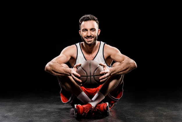 Cheerful basketteur assis avec ballon sur fond noir