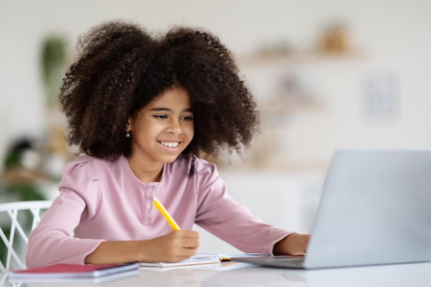 Cheerful black girl schooler faisant ses devoirs à l'aide d'un ordinateur portable