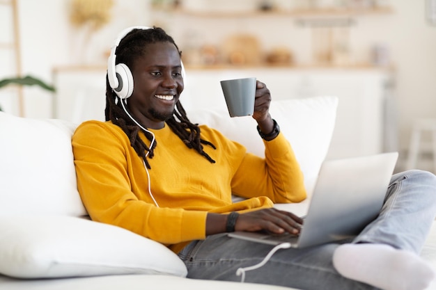 Cheerful Black Man Wearing Headphones se détendre à la maison avec ordinateur portable et café