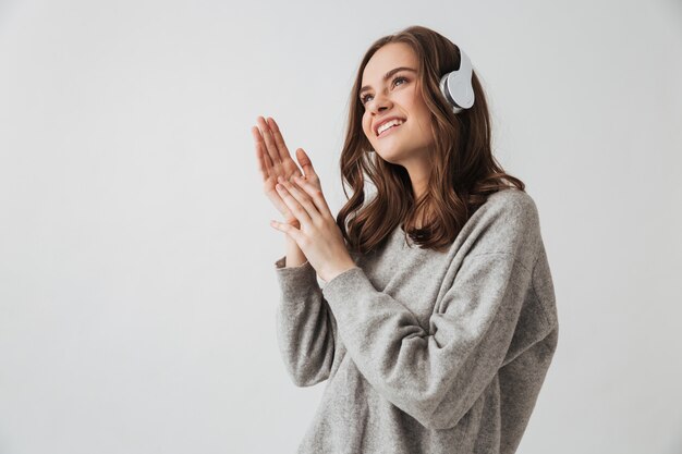 Cheerful brunette woman en pull et casque d'écoute de la musique et à la recherche sur mur gris