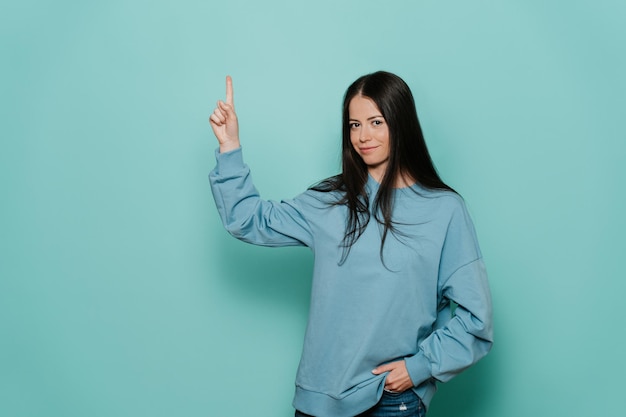 Cheerful brunette young woman in casual smiles largement pointe vers le haut à l'espace libre sur turquoise